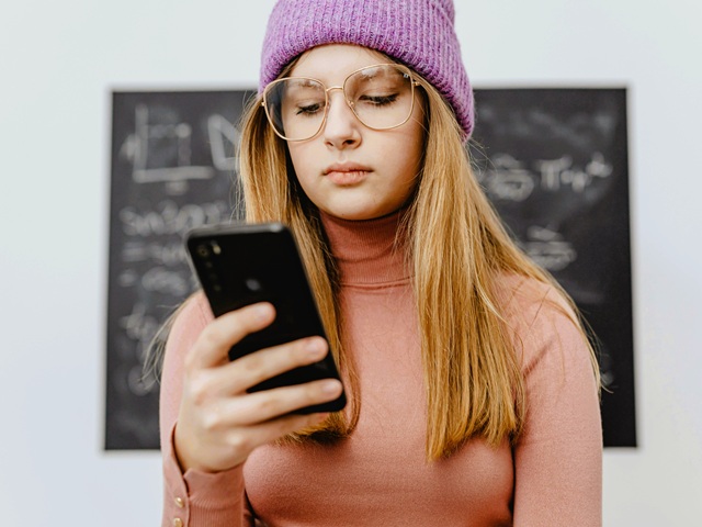Ein junges Mädchen sitzt in der Schule vor einer Tafel und schaut auf ein Smartphone.