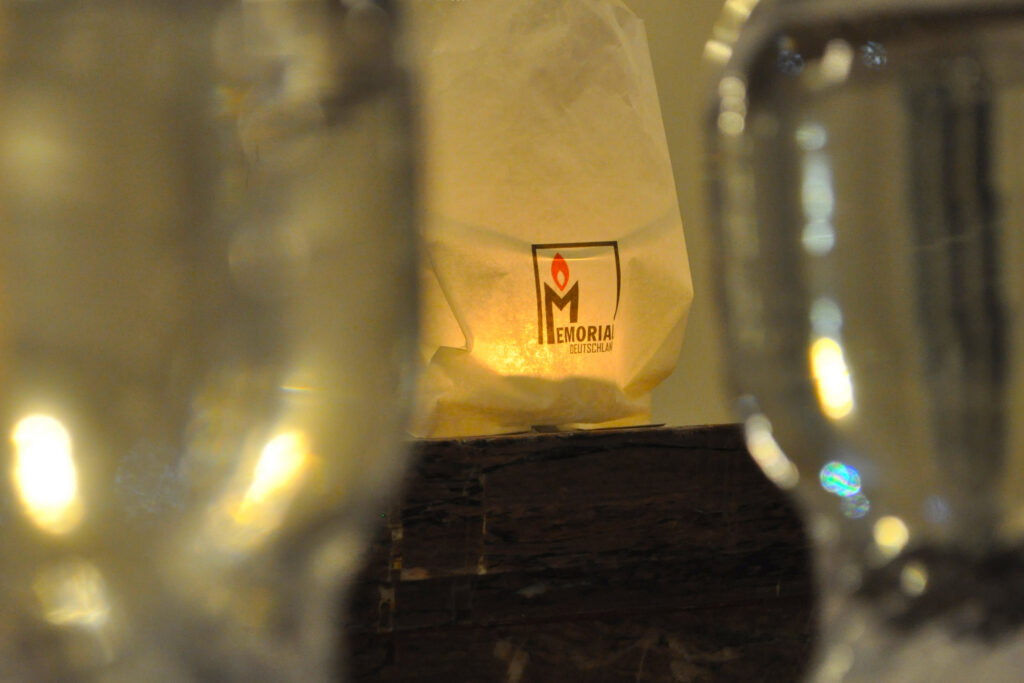 Eine Tüte mit dem Logo von Memorial Deutschland steht auf einem Tisch und funtkioniert als Kerzenhalter. Daneben stehen in Nahaufnahme Gläser.
